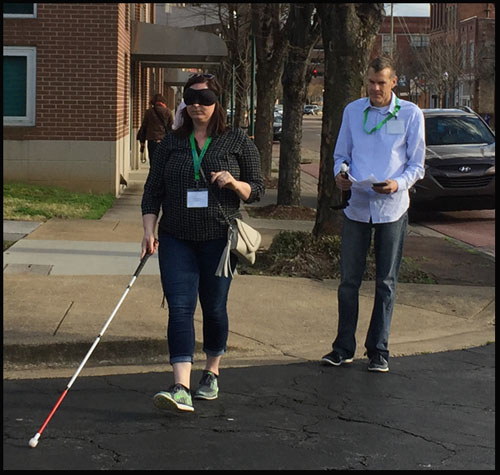 Photo shows a woman with a blindfold crossing a street while a man behind her carries his instructions.  Both are wearing green lanyards with name tags from the conference.