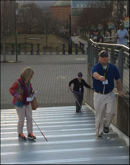 Photo shows 3 people with canes walking from a plaza up a long flight of stairs.  Two are wearing blindfolds.
