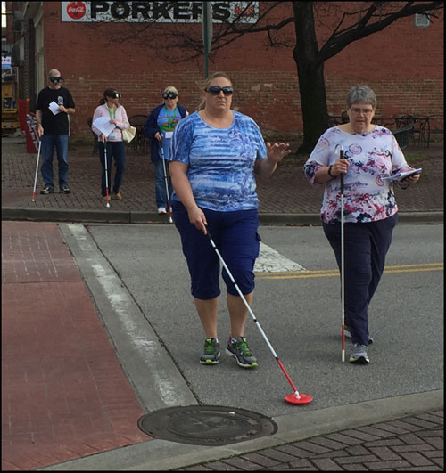 Photo shows two people crossing a narrow street with a stop sign.  One is blindfolded and using a cane.  At the corner behind them are 3 more people wearing vision simulators and using canes.