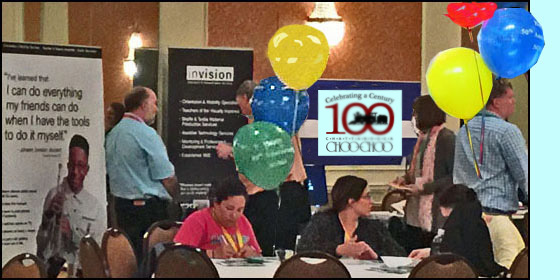 Photo shows exhibits for Invision and for Safe Toddles with people talking with the vendors.  In front of them are large round tables with balloons, and people sitting and chatting.