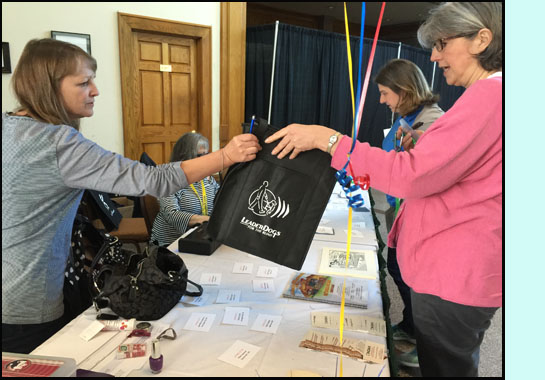 Two pictures show the table with piles of name tags and a program and bags.  Mary is sitting at her laptop and Debbie is handing people bags.
