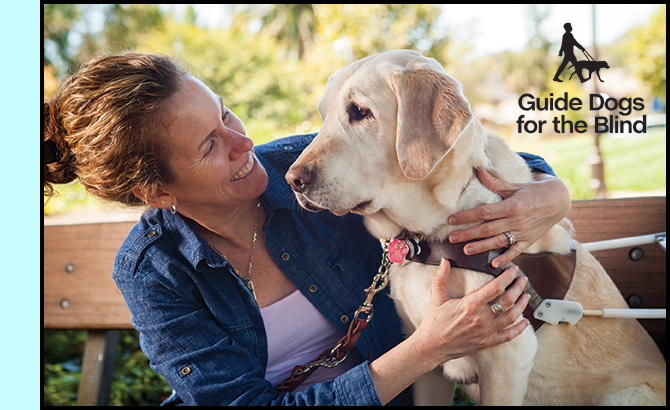 photo shows a woman sitting by a dog who is wearing a guide dog harness.  The woman is smiling broadly and looking at the dog while holding her arms around the dog's chest.