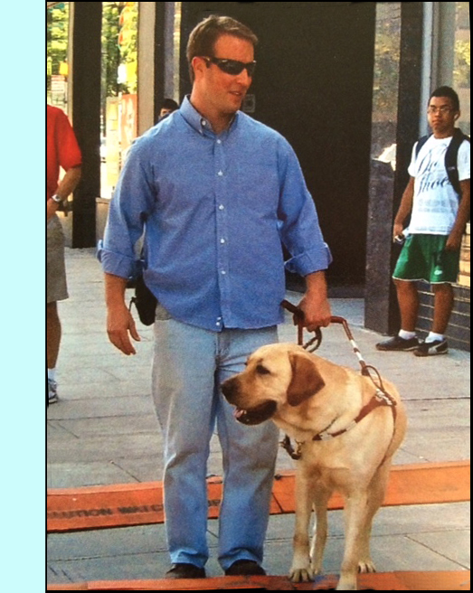 photo shows a man walking along a downtown sidewalk with a dog who is looking and seems to be turning to their right.