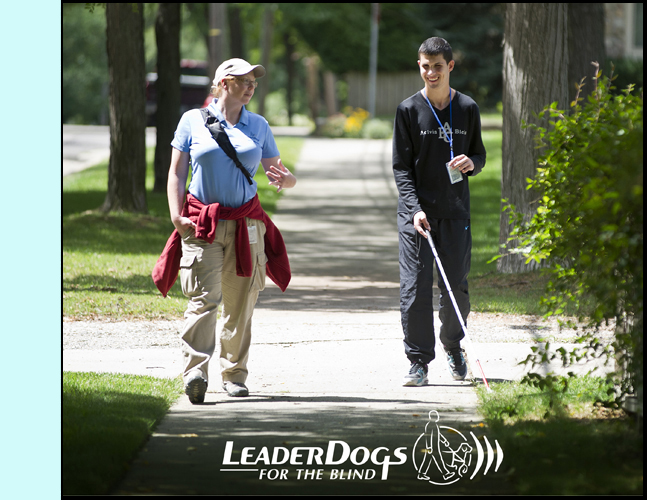 photo shows a young man walking with a cane along a sidewalk next to an instructor. She is talking to him and he is smiling.