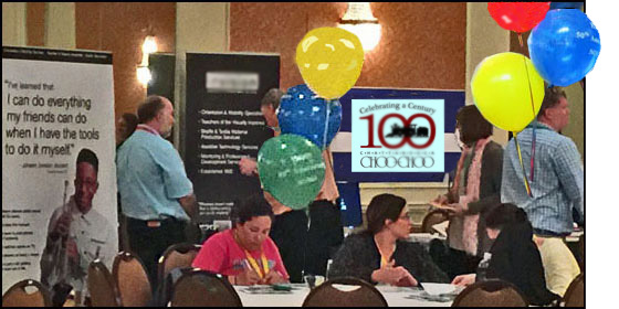 Photo shows exhibits with people talking with the vendors.  In front of them are large round tables with balloons, and people sitting and chatting.